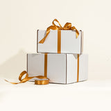 Two white gift boxes with gold ribbons stacked on a light beige background