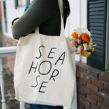 Cotton canvas Seahorse tote bag with black wordmark, worn over shoulder outdoors with flowers inside